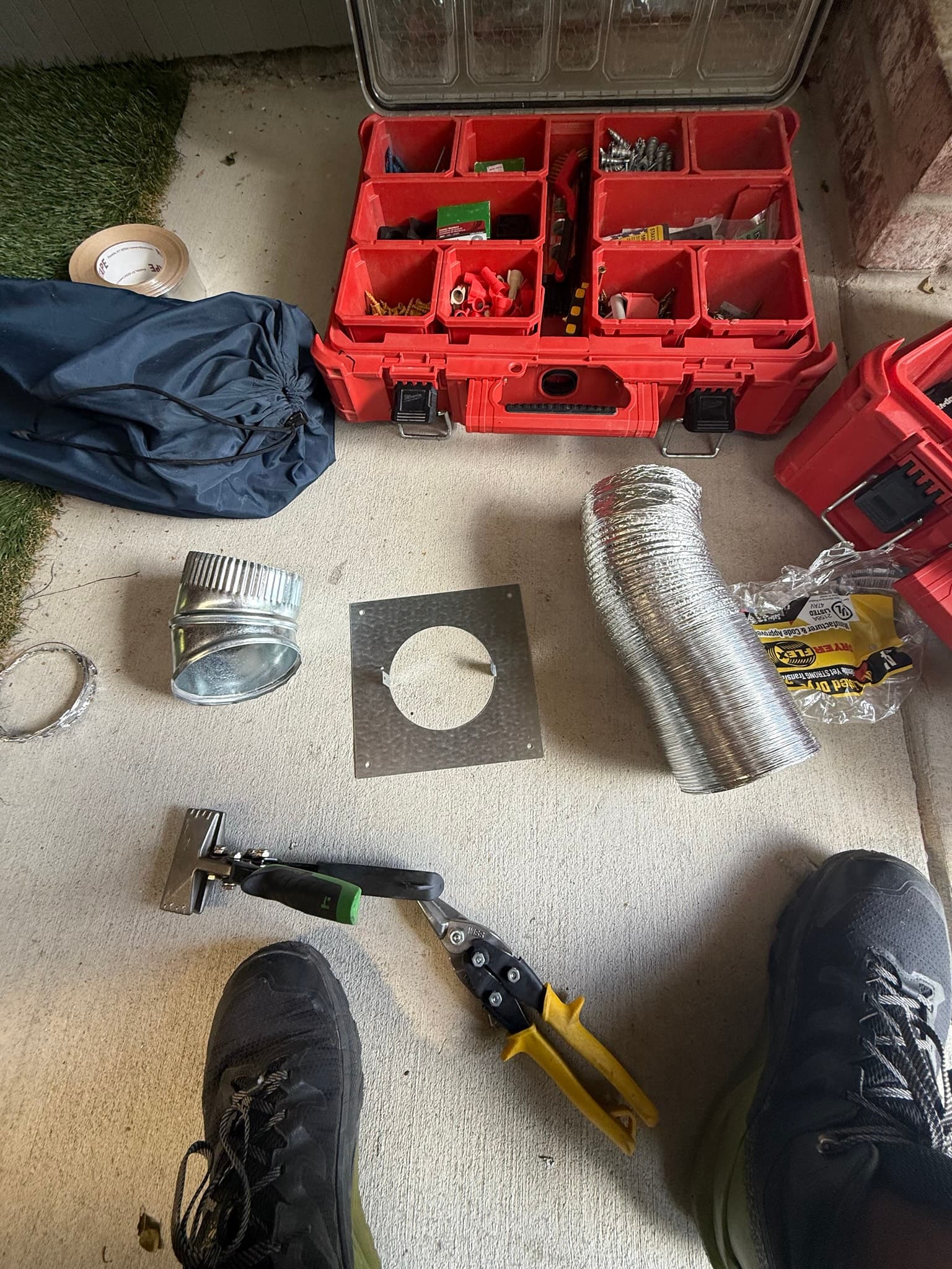 Service materials laid out on a concrete floor including a flexible aluminum dryer vent duct, metal wall flange, metal hose clamps, a putty knife, fasteners in a red organizer, and various hand tools
