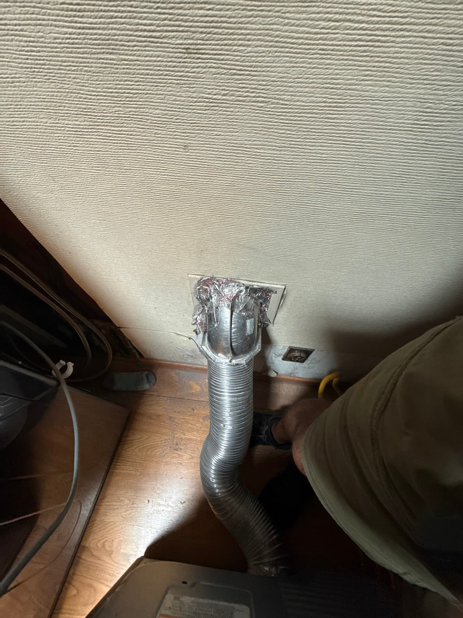 Flexible metal dryer exhaust duct connected to a wall outlet box, with the joint wrapped in clear plastic rather than secured with a proper mechanical clamp, and visible dust accumulation on the duct exterior