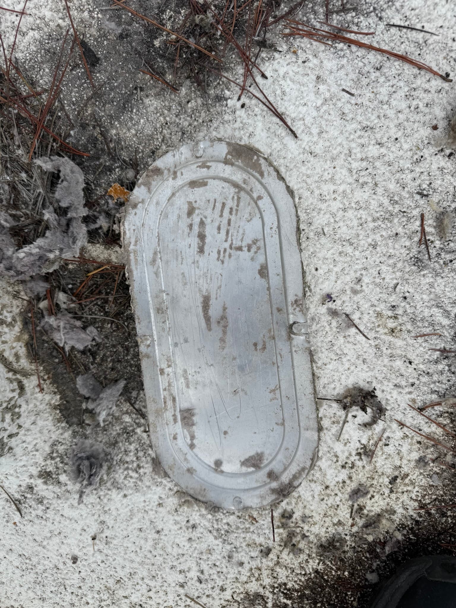 Removed exterior dryer vent cap lying on concrete surrounded by dead vegetation, its white plastic housing discolored with heavy lint, dust, and debris clogging the interior louver slots