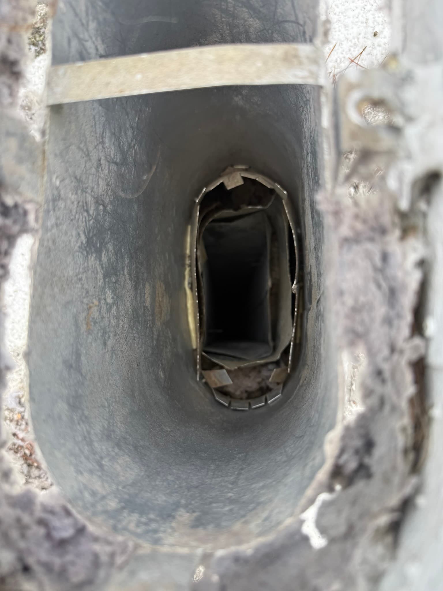 Dryer vent opening in an exterior wall with a bent or deformed flapper, heavy lint buildup on interior duct surfaces, and white and gray lint dust coating the surrounding wall area