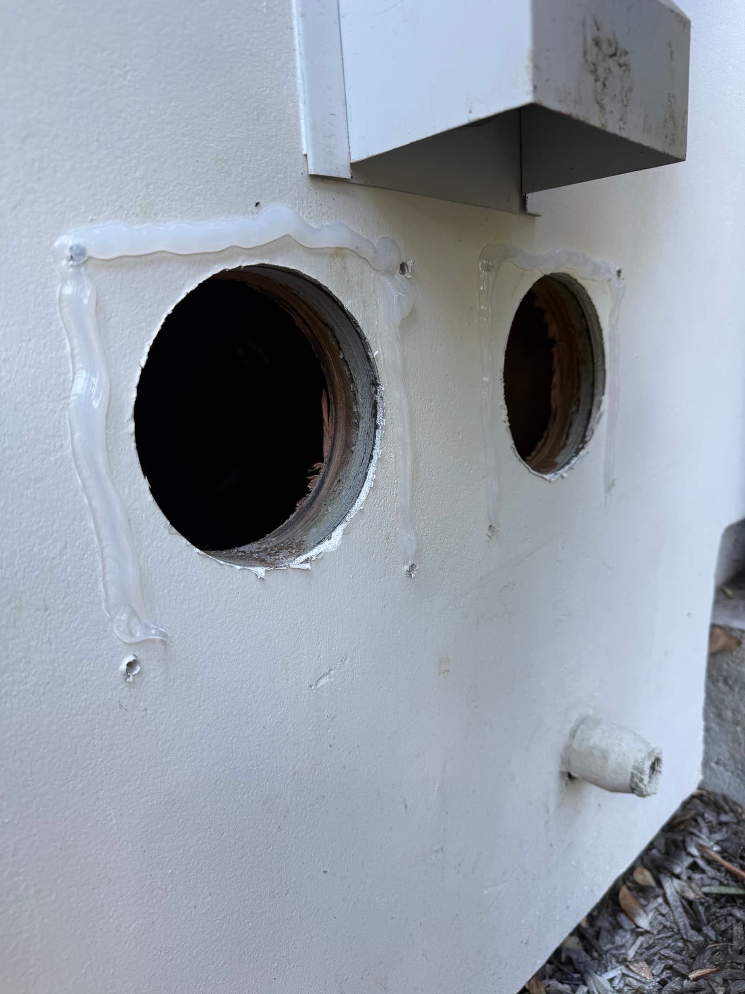 Two circular wall penetrations in exterior stucco, prepped with caulk and ready for new vent hood installation, with lint debris visible around the left opening