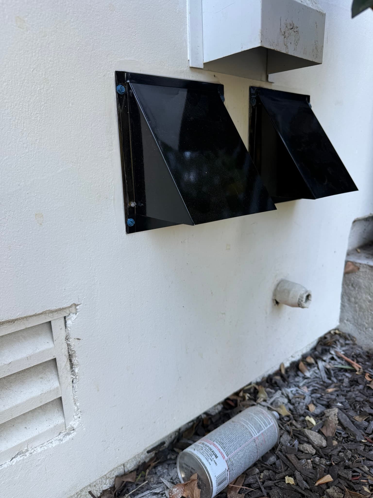 Finished exterior dryer vent hood on white stucco wall, painted black to match the home's window trim, with dual damper doors closed