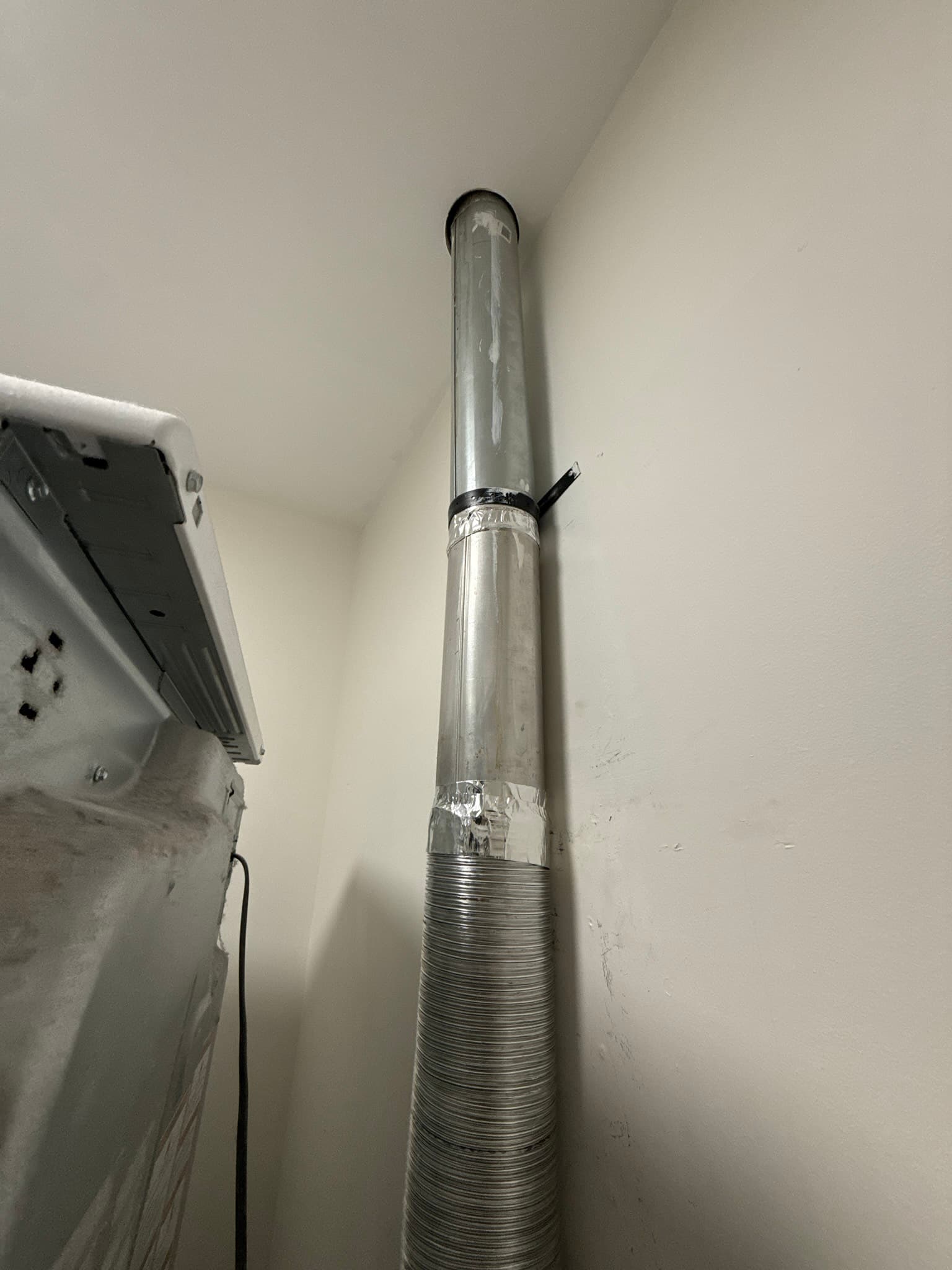 Interior view of accordion-style flexible dryer vent duct secured with a metal band clamp at the lower section, transitioning to a rigid metal pipe with a black damper at the top, with a white dryer visible at the left