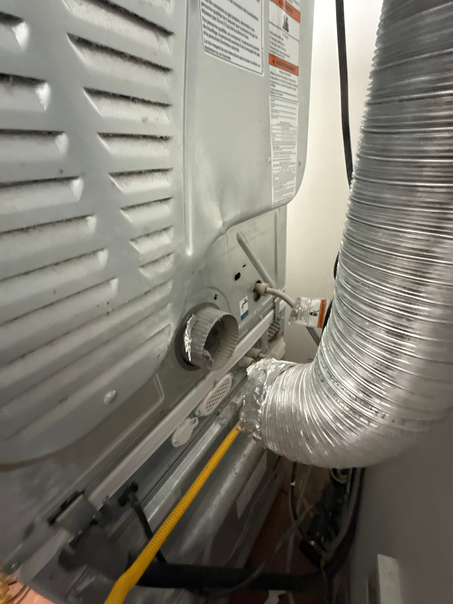 Rear of dryer unit showing the new flexible aluminum DryerFlex transition duct connected to the dryer outlet, with a yellow gas line running alongside on the floor