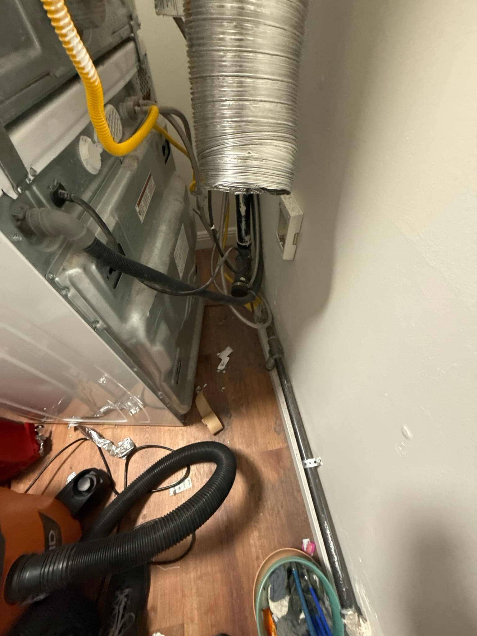 Dryer installation showing a yellow flexible duct at the dryer outlet and a silver semi-rigid metal duct extending upward toward the wall, with black flexible duct visible on the floor and a loose or improperly secured connection