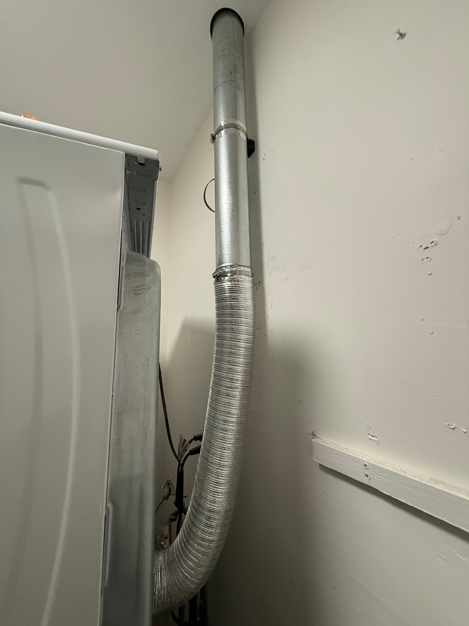 Dryer vent transition assembly showing a rigid metal duct in the upper section connected via hose clamps to a flexible aluminum foil duct below, with visible dust and lint accumulation on the exterior surfaces
