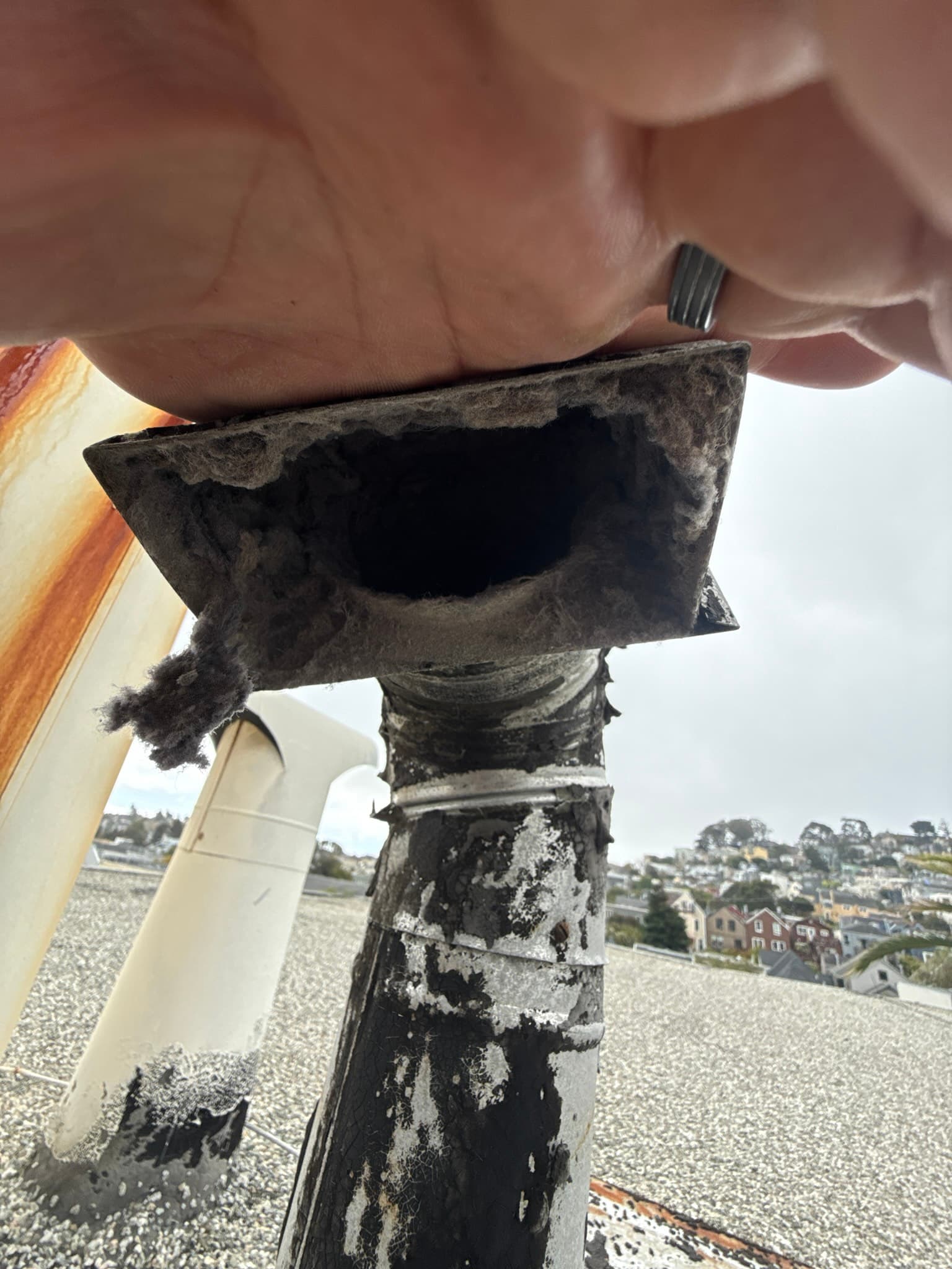 Exterior dryer vent wall cap heavily clogged with compacted lint and debris blocking the opening, with the metal duct connector below showing rust, corrosion, and white efflorescence from moisture exposure