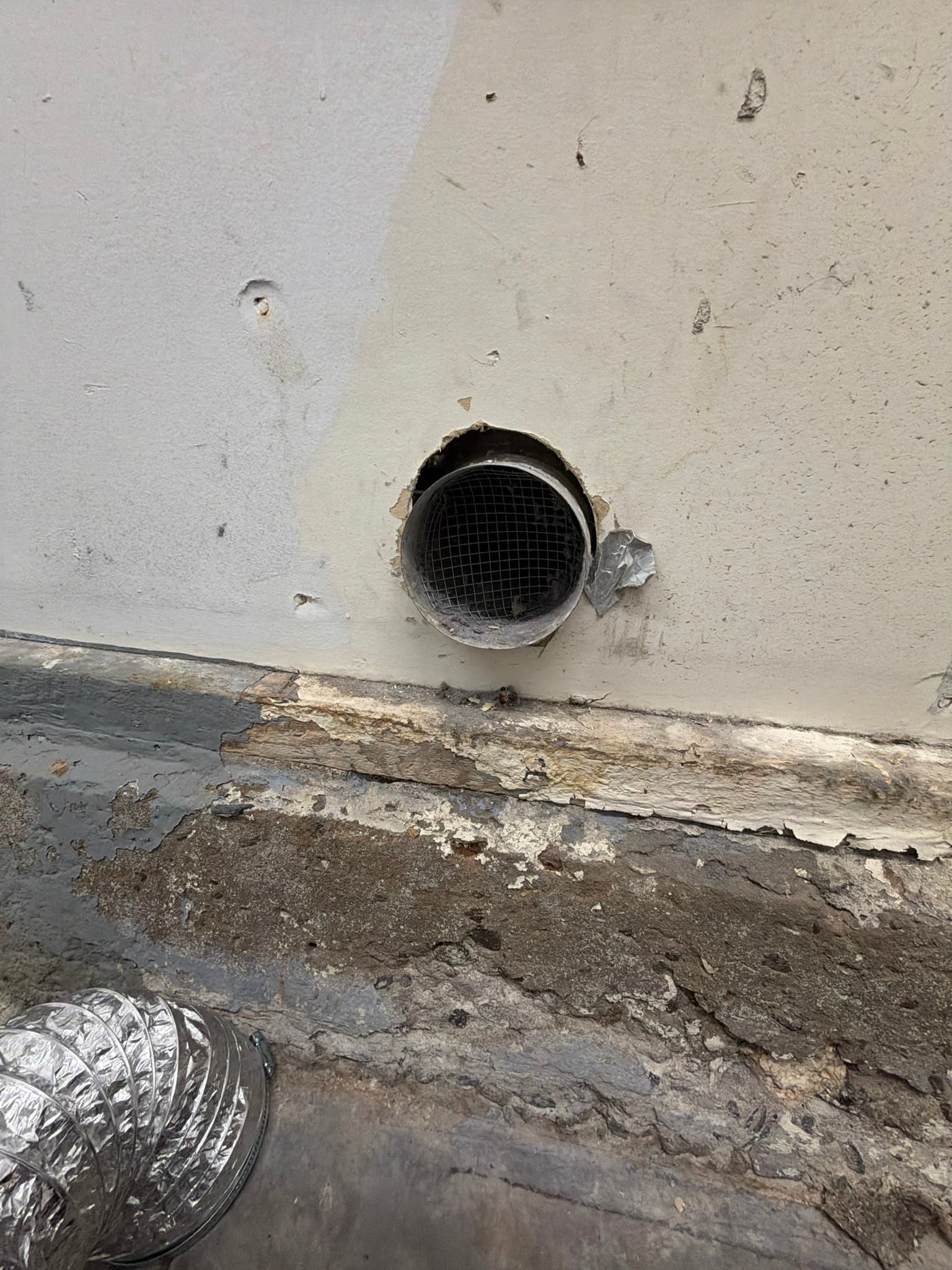 Exterior wall penetration with a metal duct covered only by a wire mesh screen instead of a proper louvered cap, with peeling foil tape and a coil of new flexible duct staged on the ground nearby