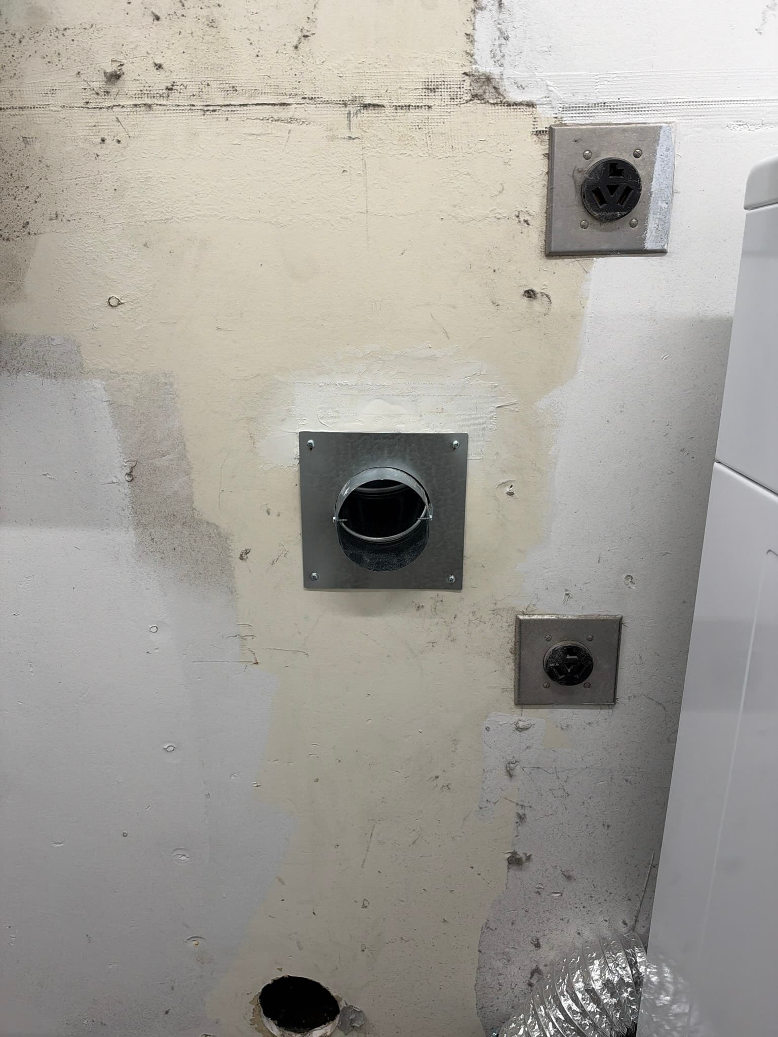 Newly installed 4 inch metal wall vent collar secured into patched drywall, with flexible transition duct connected behind it via a hose clamp and foil flex duct visible at the dryer connection