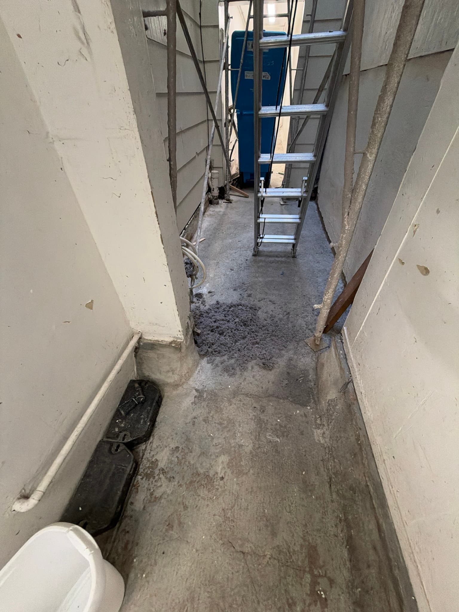 Pile of lint accumulated on a concrete walkway near a floor drain in a narrow side yard, with scaffolding and a ladder staged for access to the exhaust duct above