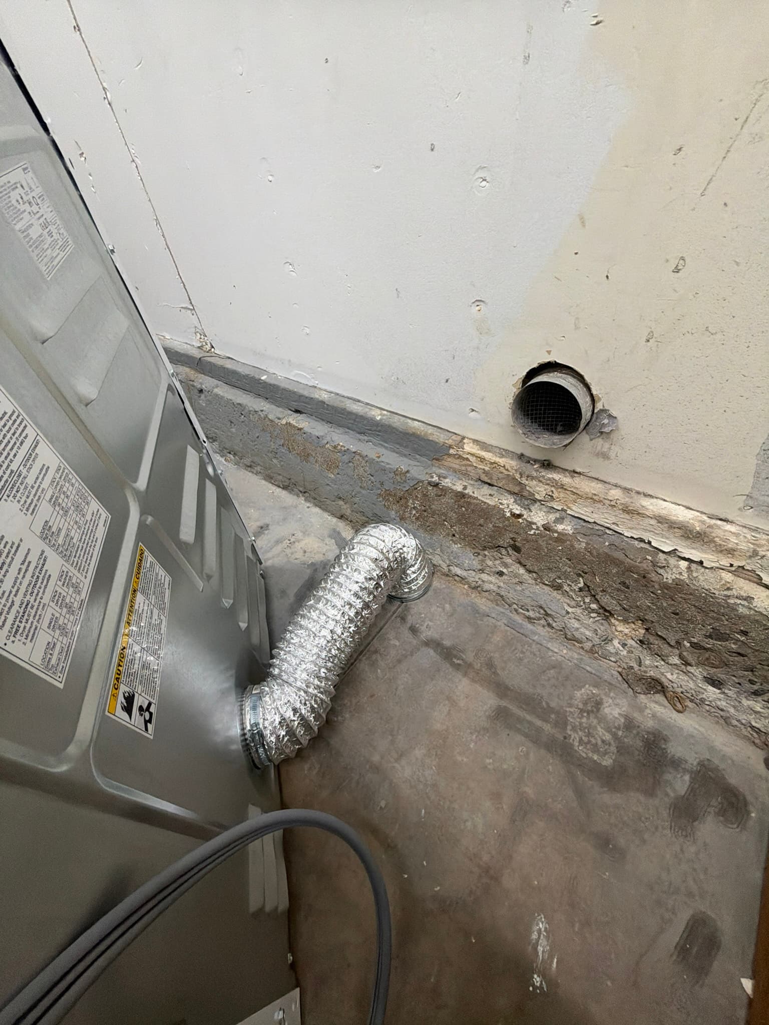 Dryer's rear panel connected to a short length of flexible foil transition duct routed to a nearby wall termination point with a bare metal collar visible through the wall opening