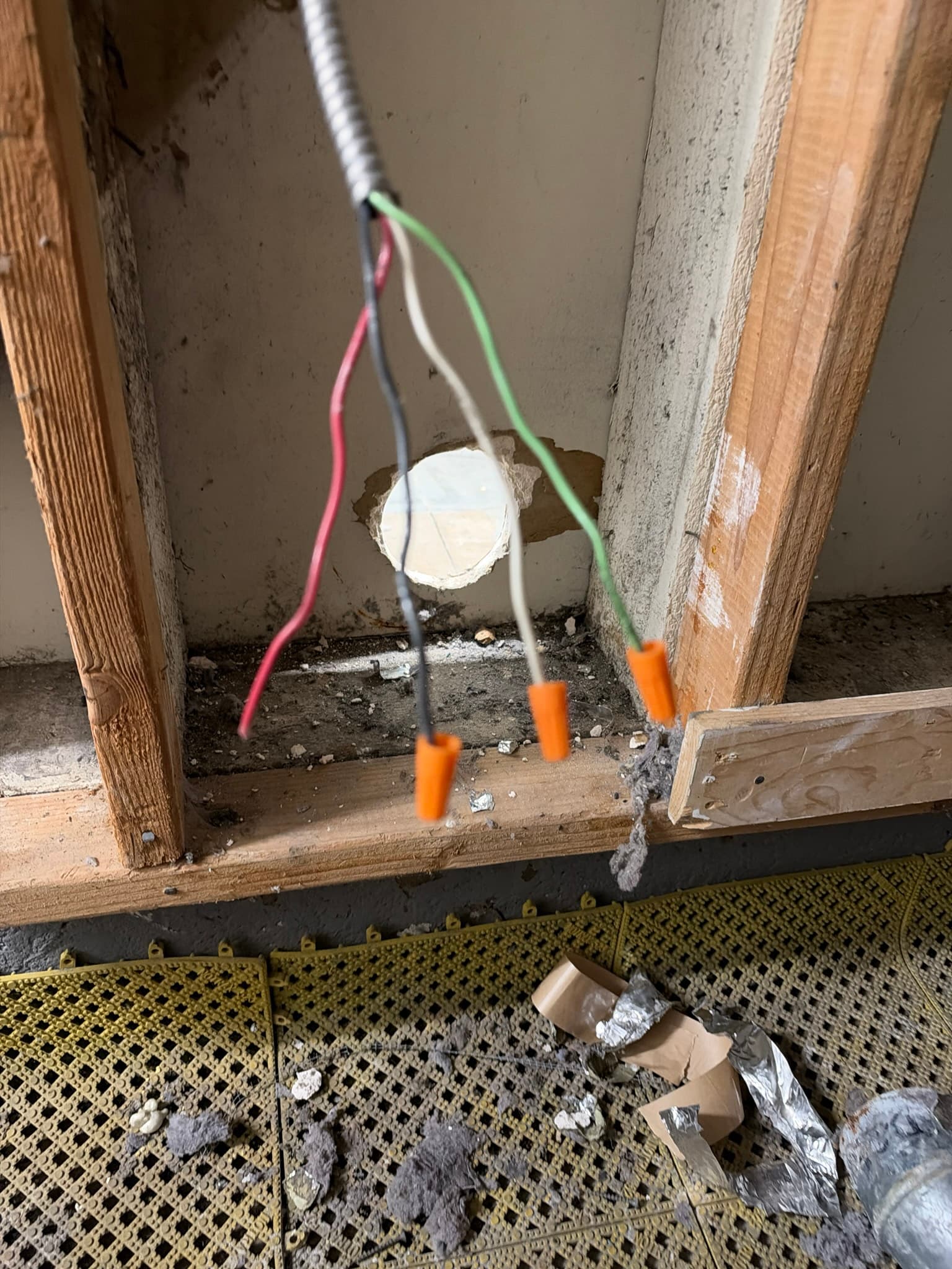 Open wall cavity with exposed electrical wiring near a cut-out access hole and scraps of lint-covered foil duct on the floor, left over from removing the old transition duct
