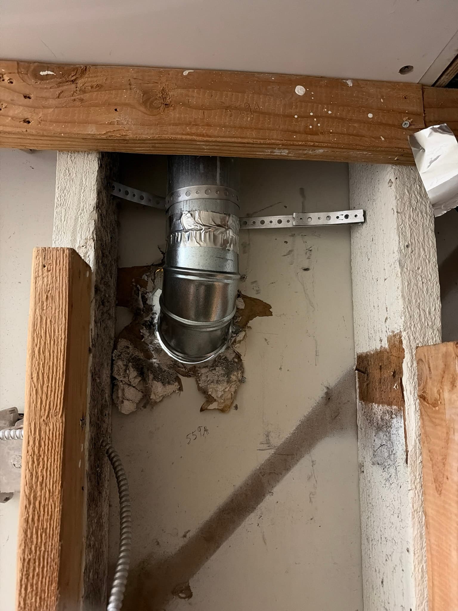 Open wall cavity showing rigid metal duct sections joined with foil tape and screws, secured to framing with a perforated metal strap, with deteriorated insulation nearby