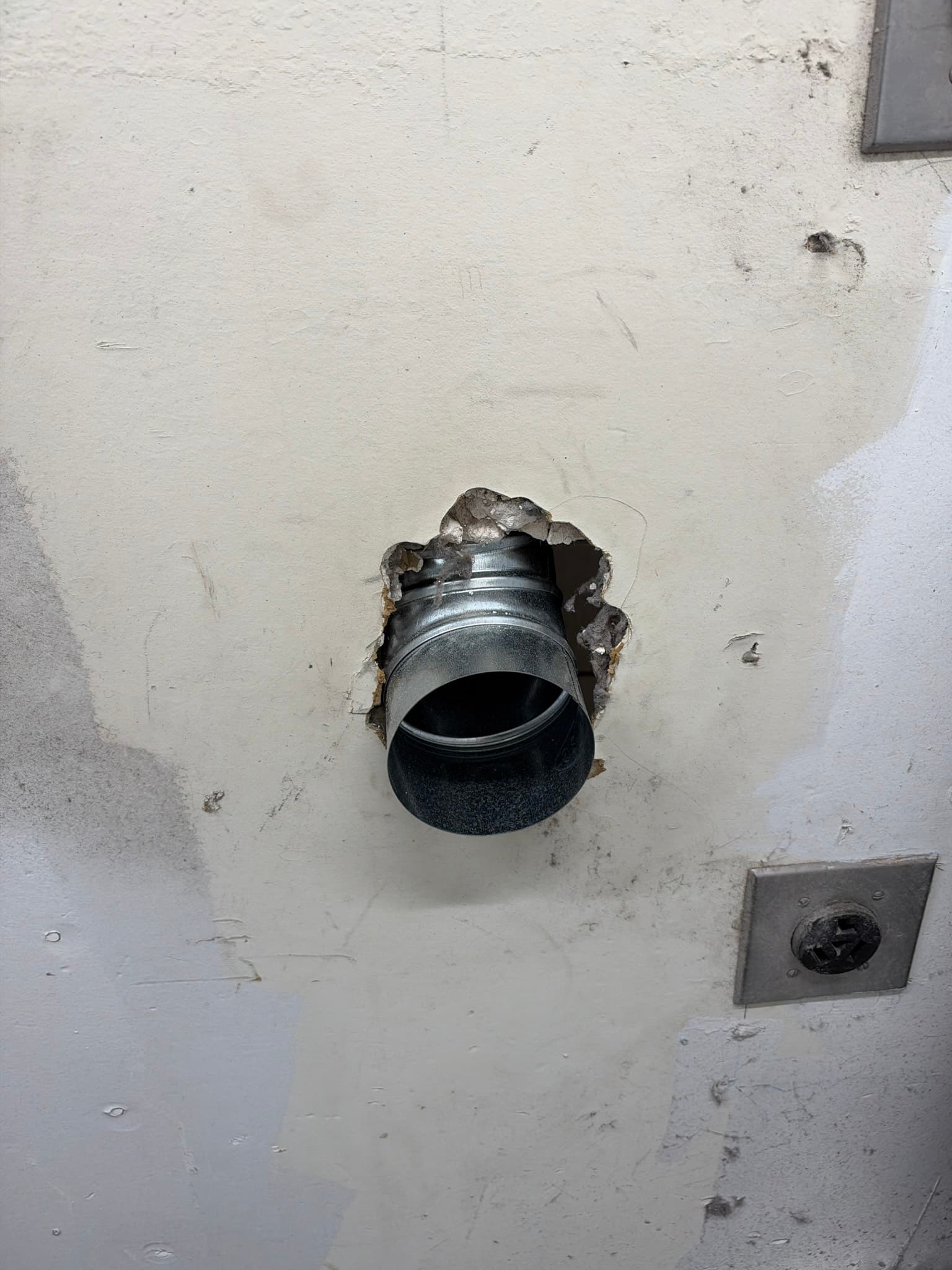 Metal transition duct protruding through a roughly broken interior wall opening with torn drywall and debris around the penetration prior to repair