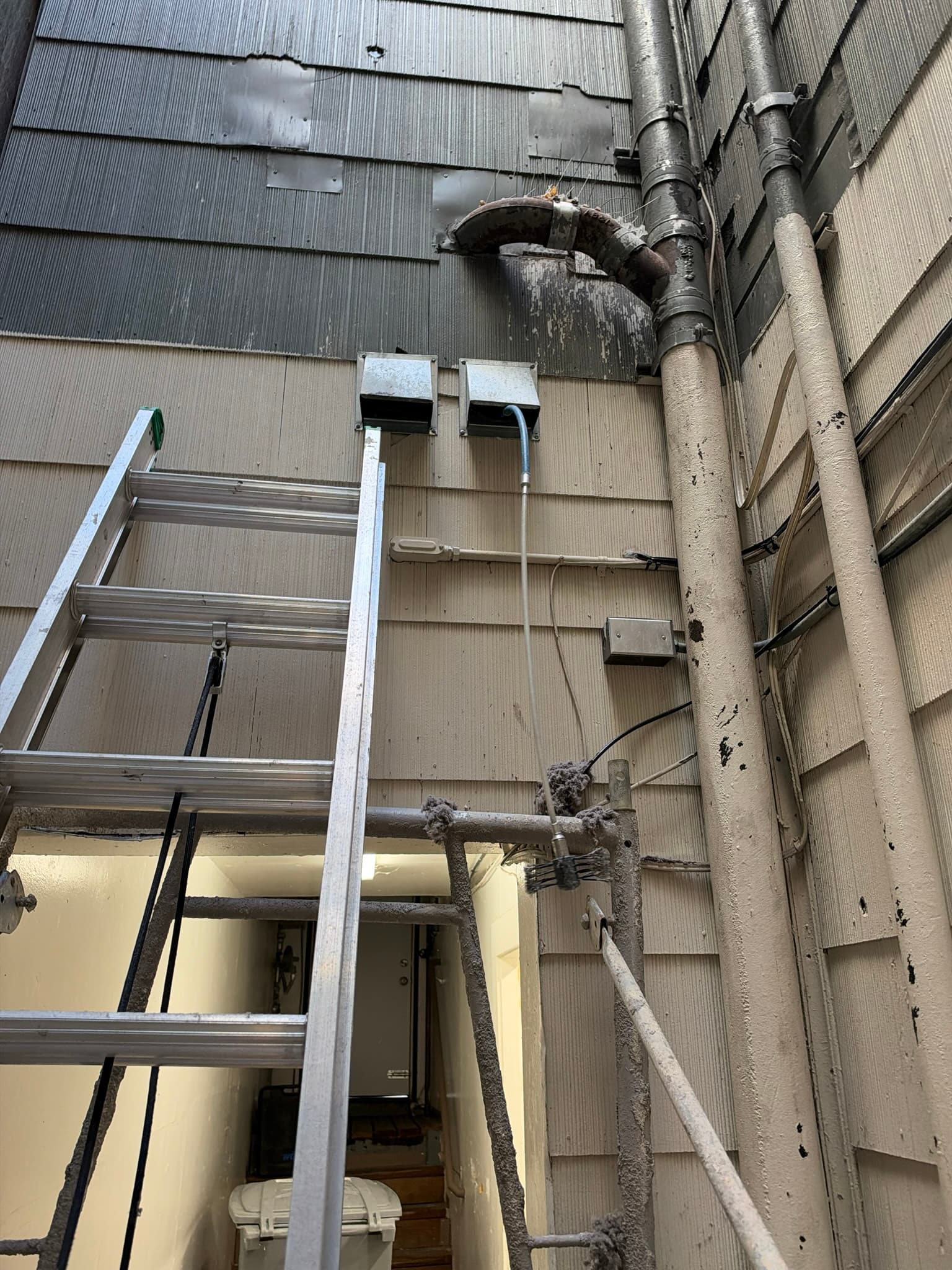 Corroded metal elbow fitting on the roofline packed with heavy lint buildup, with additional lint visible on metal piping lower on the exterior wall near a ladder set up for access