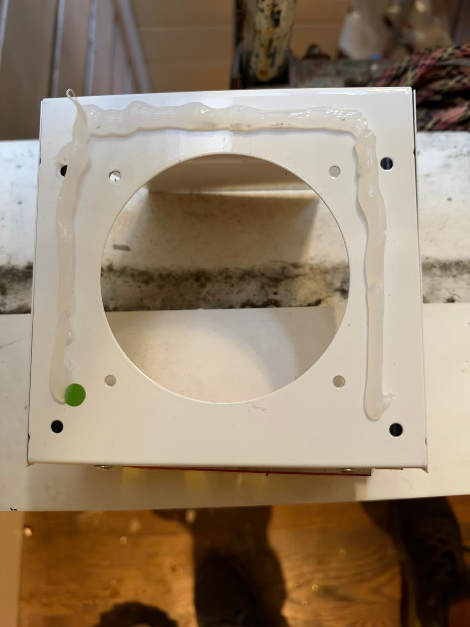 Back of the white flush mount dryer wall vent showing a bead of sealant applied around the mounting flange and pre-drilled mounting holes around the duct opening