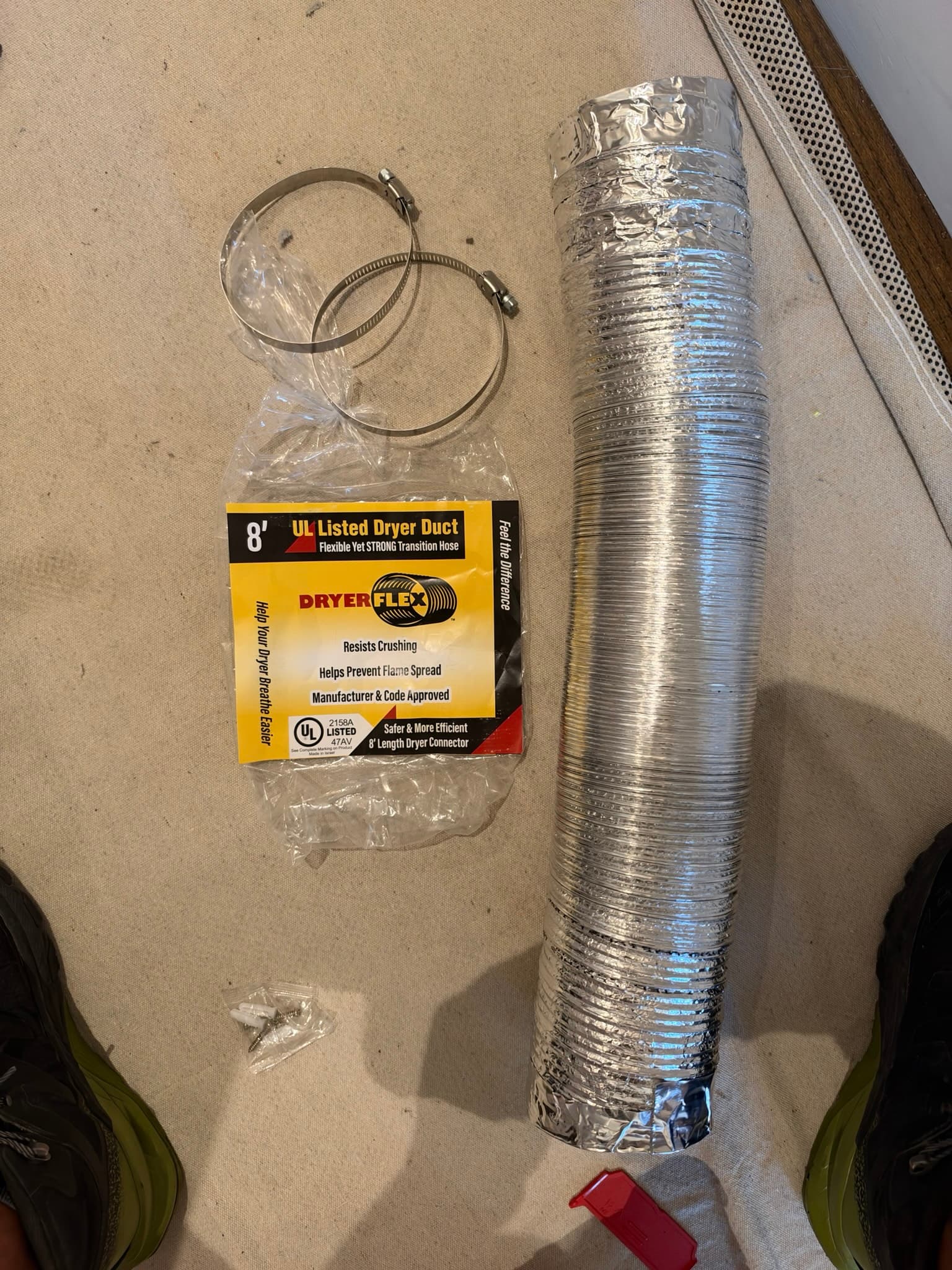 Coiled 8-foot DryerFlex transition duct in UL listed packaging alongside two stainless steel worm-gear clamps and mounting screws, staged before installation