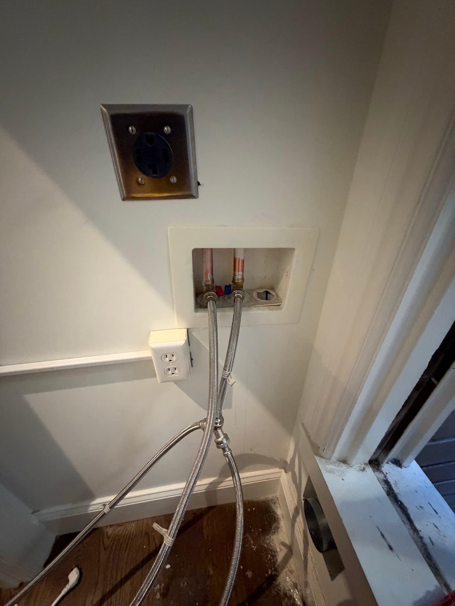 Laundry alcove showing a 240V dryer outlet, washer supply box with braided hoses, duplex outlet, and a dusty flush-mounted dryer vent opening in the wall